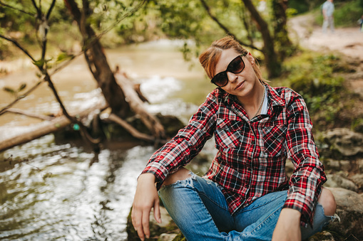One young woman in plaid shirt and sunglasses sitting by the river in nature and enjoying free time on sunny spring day