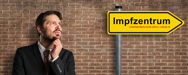 street sign with German message for VACCINATION CENTER and a thinking businessman in front of a brick wall background
