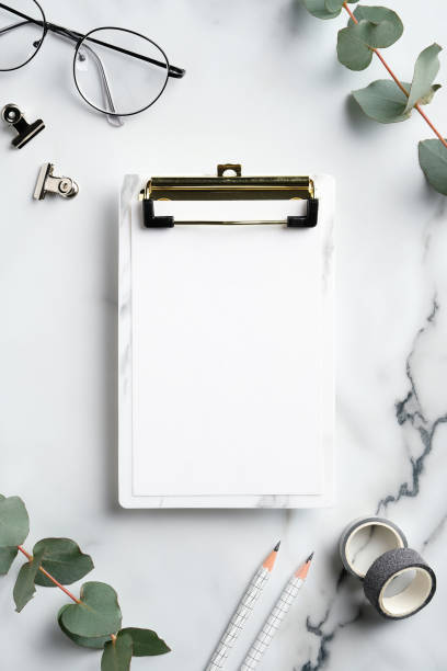 istock Home office workspace with clipboard mockup, glasses, supplies, greenery on marble table. Flat lay, top view. 1298998994