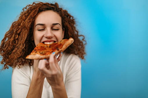 Woman standing in front of blue background and eating pizza