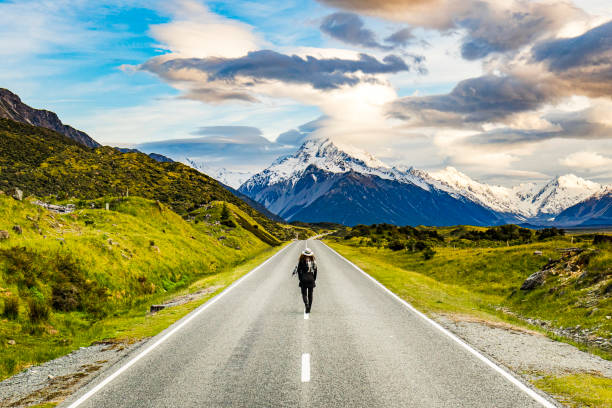istock Young adventurous woman walking down road towards large snowy mountain 1299015636