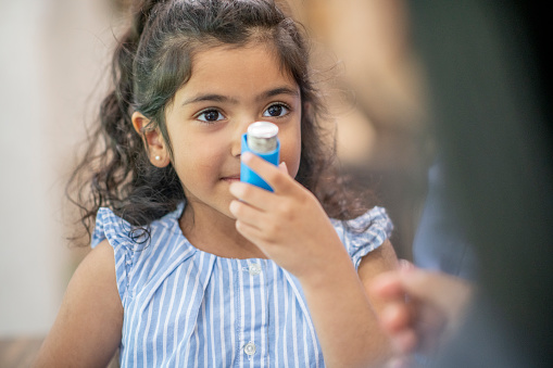 A young girl of Hispanic ethnicity is holding an asthma inhaler close to her mouth to demonstrate how she uses it.