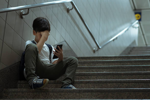 Asian teenager boy sitting alone at staircase, covering his face with his hand, holding smartphone, crying, feeling depressed, stressed due to cyberbullying at school. Social issues in internet technology and social media in teenager mental health. Low self esteem young Asian teenager boy sitting alone crying with smartphone, feeling frustration, fear, pain, anxiety, abused as victim of cyberbullying. cyberbullying stock pictures, royalty-free photos & images