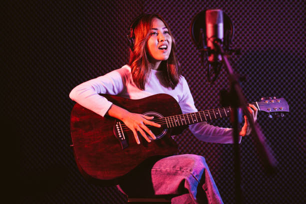 istock Pretty Asian female singer recording songs by using a studio microphone and pop shield on mic while playing guitar and wearing headphones in the studio. Performance and show in the music business. 1299034705