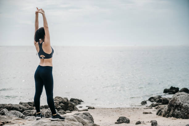istock Asian fitness runner body closeup doing warm-up routine on beach before running, stretching leg muscles with standing single knee to chest stretch. Female athlete preparing legs for cardio workout. 1299036285