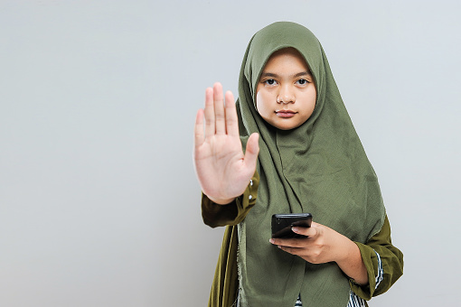 Young Muslim woman holding mobile phone with stop bullying gesture, isolated on gray