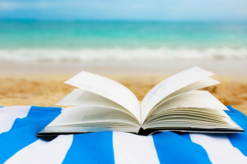 A book opened lying on a blue towel on the sand in front of a tropical ocean.