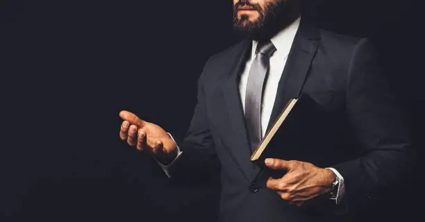 Photo of a man in a suit holding a bible in his arm speaking to another person on a black background