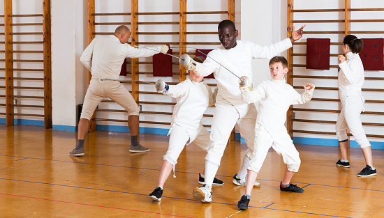 Trainer demonstrating to young athletes stances and movements with rapier during fencing workout in gym