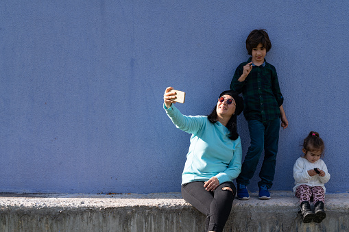 Photo of mother taking selfie photo with her 8 years old son and 2,5 years old daughter in outdoor. The background is blue colored wall. Shot in outdoor with a full frame mirrorless camera.