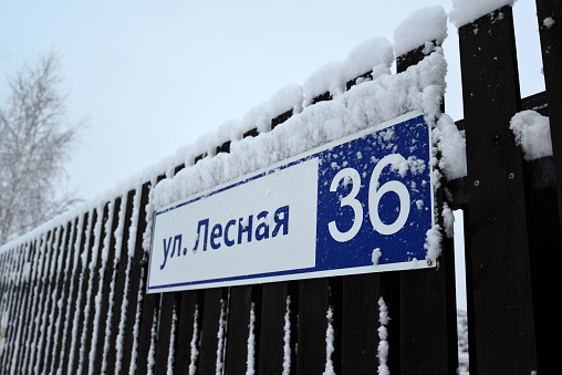 A fragment of a dark fence with a sign indicating the name of the street: Lesnaya, thirty-six. Everything is covered with snow and frost.