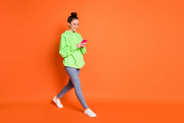 Full length photo portrait of woman texting holding phone in two hands walking isolated on vivid orange colored background Full length photo portrait of woman texting holding phone in two hands walking isolated on vivid orange colored background. topknot stock pictures, royalty-free photos & images