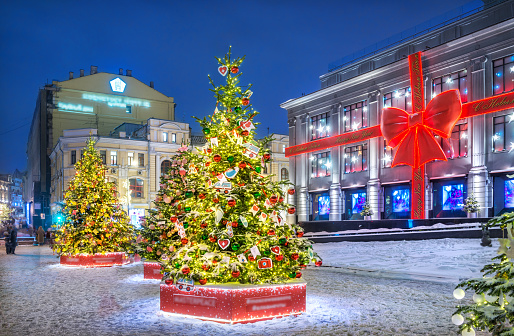 New Year trees on the square in front of the Central Department Store in Moscow and decoration on the storefront in the form of a red bow in the light of evening lanterns. Caption: Happy New Year!
