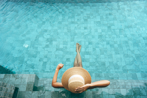 Summer domestic in Thailand travel on holiday concept. Top view of asian woman relax at swimming pool spa resort. She sitting on poolside with straw hat and bikini.
