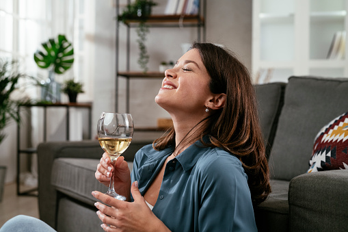 Beautiful woman sitting on the floor, drinking wine. Young woman celebrate with wine at home.