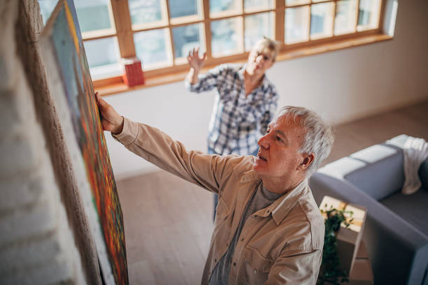 senior couple in their new home hanging a painting together - home decorating fotos imagens e fotografias de stock