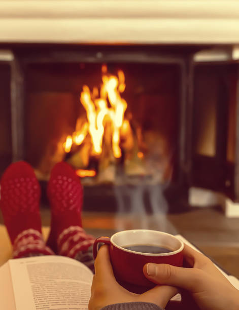 Person resting in front of the fireplace with a steaming hot cup of coffee and a book. Shallow depth of field. Person resting in front of the fireplace with a steaming hot cup of coffee and a book. Shallow depth of field. heat home interior comfortable human foot stock pictures, royalty-free photos & images