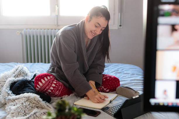 Young caucasian woman writing in a notebook above the bed Young Caucasian woman writing in a notebook above the bed in her room with pijáma clothes and with a tablet and a mobile phone on top of the bed confinamiento stock pictures, royalty-free photos & images