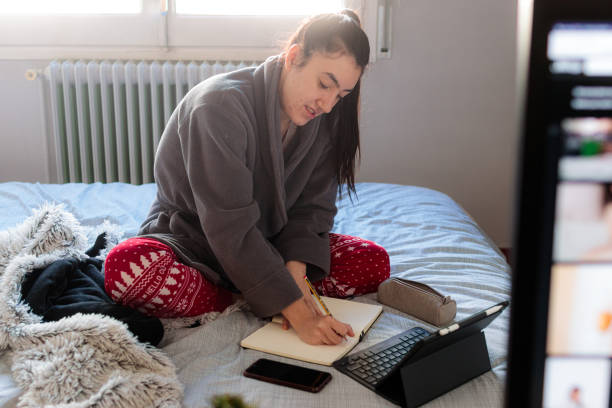 Young caucasian woman writing in a notebook above the bed Young Caucasian woman writing in a notebook above the bed in her room with pijáma clothes and with a tablet and a mobile phone on top of the bed confinamiento stock pictures, royalty-free photos & images