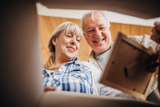 Senior couple unpacking cardboard box together Two people, senior couple opening cardboard box after moving in new home and looking at framed picture. seniors-moving-house stock pictures, royalty-free photos & images