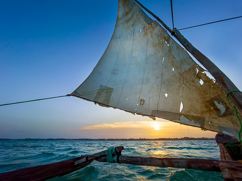 Old traditional maritime traditional vessel Dhow boat sailing under torned sail in the open Indian ocean near Zanzibar island in beautiful sunset, Tanzania. Traveling and unique local culture concept.