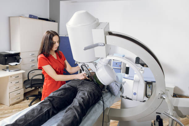 istock Extracorporeal Shock Wave Lithotripsy. Young female doctor in red uniform performing lithotripsy procedure for male patient, using modern equipment, ultrasonic lithotripter to break up kidney stones 1299086866