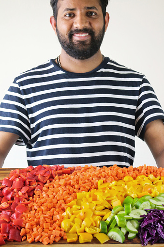 Stock photo showing a close-up of Indian man holding chopping board with rainbow display of multi-coloured chopped fruit and vegetables including shredded red cabbage, sliced cucumber, yellow pepper, diced carrot and tomato, elevated view, healthy eating concept
