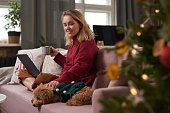 Happy young woman with blond hair wearing stylish warm outfit spending Christmas morning at home sitting on sofa, petting her lovely dog and using laptop