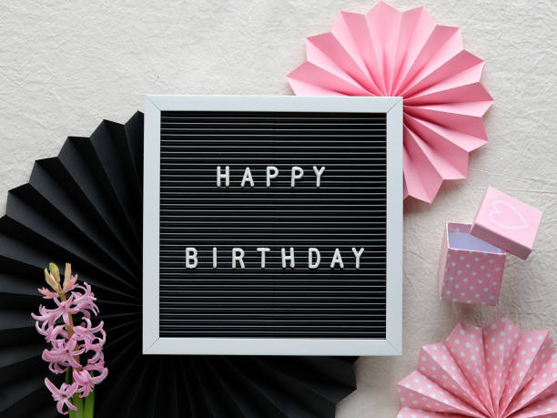 istock Text Happy Birthday on letter board, letterboard. Creative flat lay in pink and black on ivory, cream white textile. Pink hyacinth flowers. Party decor, paper fans, sugar hearts, perfume glass bottle 1299096127