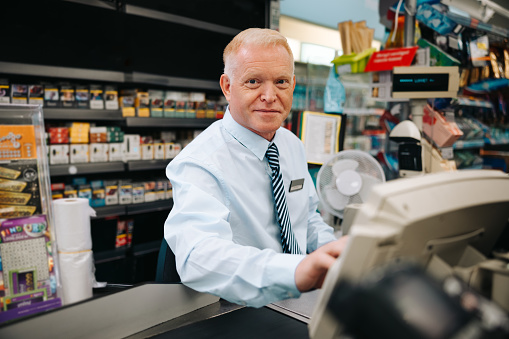 Senior man sitting at super market checkout and looking at camera. Grocery store manager at checkout counter.