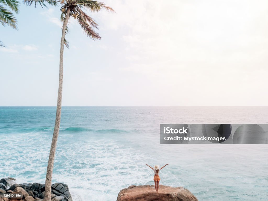 Woman on rock above sea stands arms out wide Young woman on palm tree hill standing on rock arms wide open embracing nature freedom and life. People balance spirituality healing outdoors Aura Stock Photo
