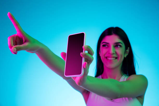 istock Brunette beautiful woman's portrait isolated on blue studio background in multicolored neon light. Showing blank phone's screen 1299115116