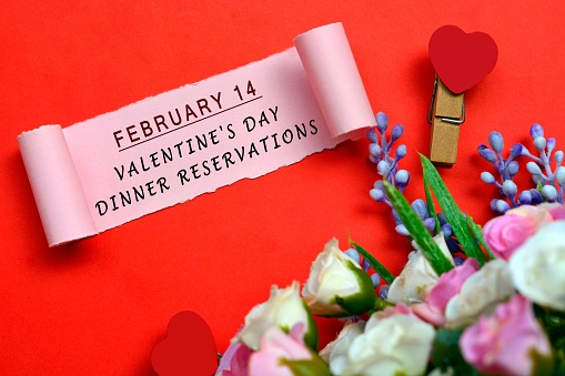 February 14 Valentine's day dinner reservations label on torn paper with flowers and heart shape clothespin on red background. Valentine's Day Concept