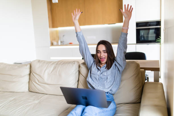 istock Pretty young woman using a laptop while raising hand win gesture and sitting on the couch 1299122010