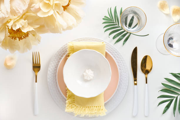 istock Effortless golden birthday dinner table decor. Pale yellow peony flowers. White dinner table, white and gold utensils, decorated with season flowers and palm leaves 1299126566