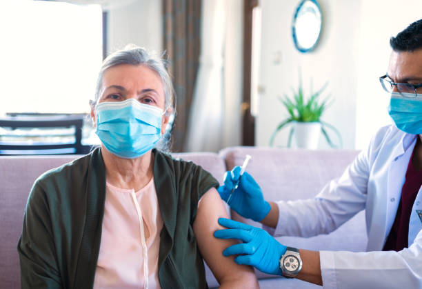 männliche krankenschwester mit schützender gesichtsmaske beim sitzen zu hause und injektion einer seniorin während corona-ausbruch. senior-patient wird zu hause geimpft. coronavirus-impfung - isolated cold virus head and shoulders cold and flu stock-fotos und bilder