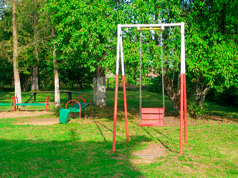 Old hanging swing of red color on a metal chain. The playground is located on green grass near large trees.