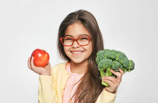 Photo of Smiling mixed race girl holding an apple and broccoli in her hands. Healthy vegetarian food for kids