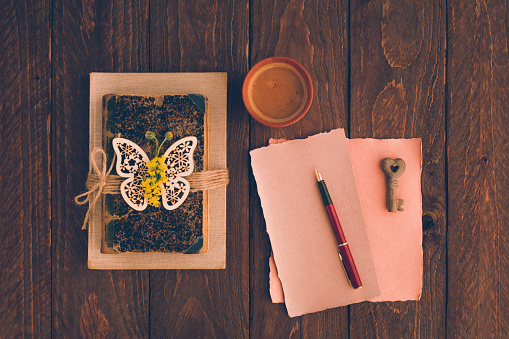 Top view of a brown flat lay in retro style with old vintage books, a wooden butterfly and flowers with a blank handmade and rustic paper mockup and a cup of creamy black coffee. Useful for wisdom, curiosity, philosophy, love and best for your soul concepts. Retro editing with added grain. Part of a series.