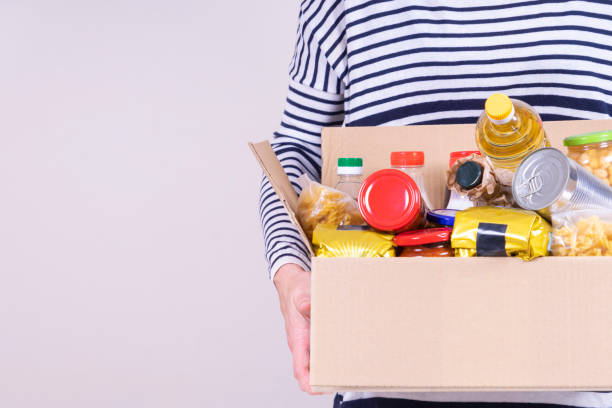 Volunteer hands holding food donations box with grocery products on white desk Volunteer hands holding food donations box with grocery products on white desk. charitable donation stock pictures, royalty-free photos & images