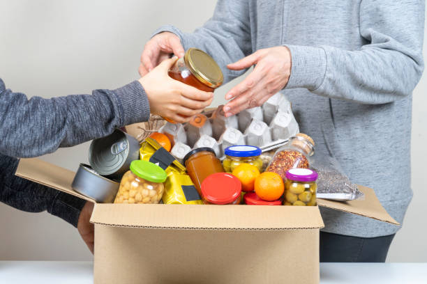 Volunteers hands putting grocery products, foodstuff to food donations box stock photo