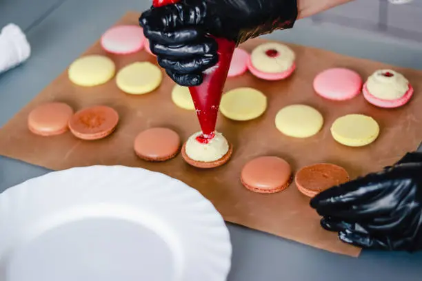 Photo of Confectioner in black rubber gloves stuffs macaroon cakes with jam