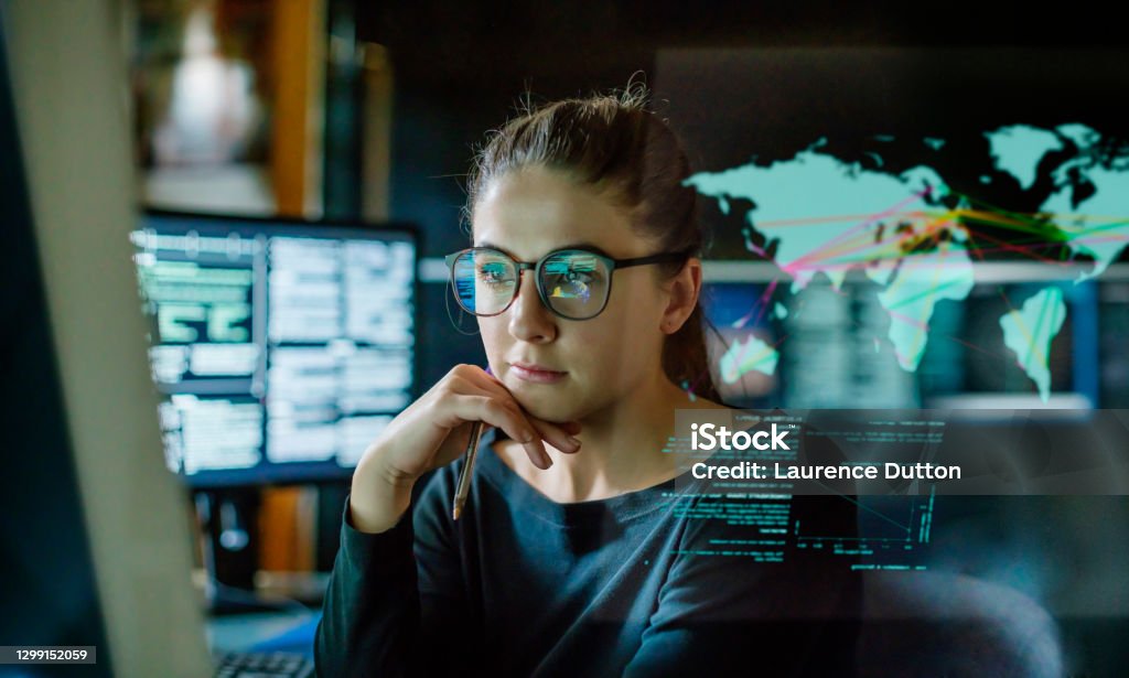 Young woman global communications Stock image of a young woman, wearing glasses, surrounded by computer monitors in a dark office. In front of her there is a see-through displaying showing a map of the world with some data. Data Stock Photo