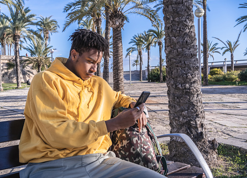 afro american black man with yellow sweatshirt looking at smartphone laying on a wooden bench in a park