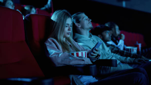 istock Beautiful young woman using her phone for texting while missing boring movie at the cinema 1299157723