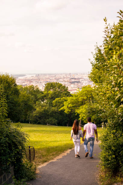 Rear view of romantic young couple holding hands and walking in the public park towards the viewpoint of the cityscape.