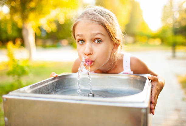 Cheerful wonderful girl drinks fresh water from a small fountain in a summer warm sunny park Cheerful wonderful girl drinks cool fresh water from a small fountain in a summer warm sunny park on a long-awaited vacation whites only drinking fountain stock pictures, royalty-free photos & images