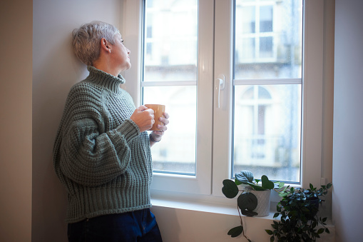 Senior woman having cup of coffee looking at the window outside from home