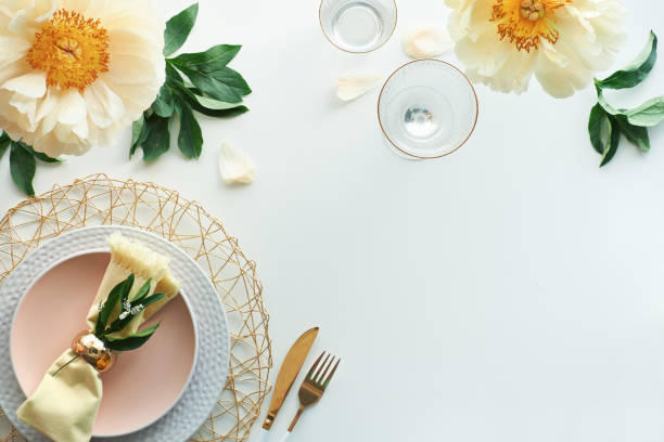 istock Effortless golden birthday dinner table decor. Pale yellow peony flowers. White dinner table, white and gold utensils, decorated with season flowers and palm leaves 1299173388