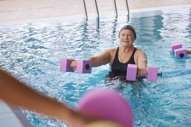 femme aîné faisant l’aérobic d’eau soulevant des haltères dans la piscine intérieure - water aerobics swimming pool exercising sport photos et images de collection